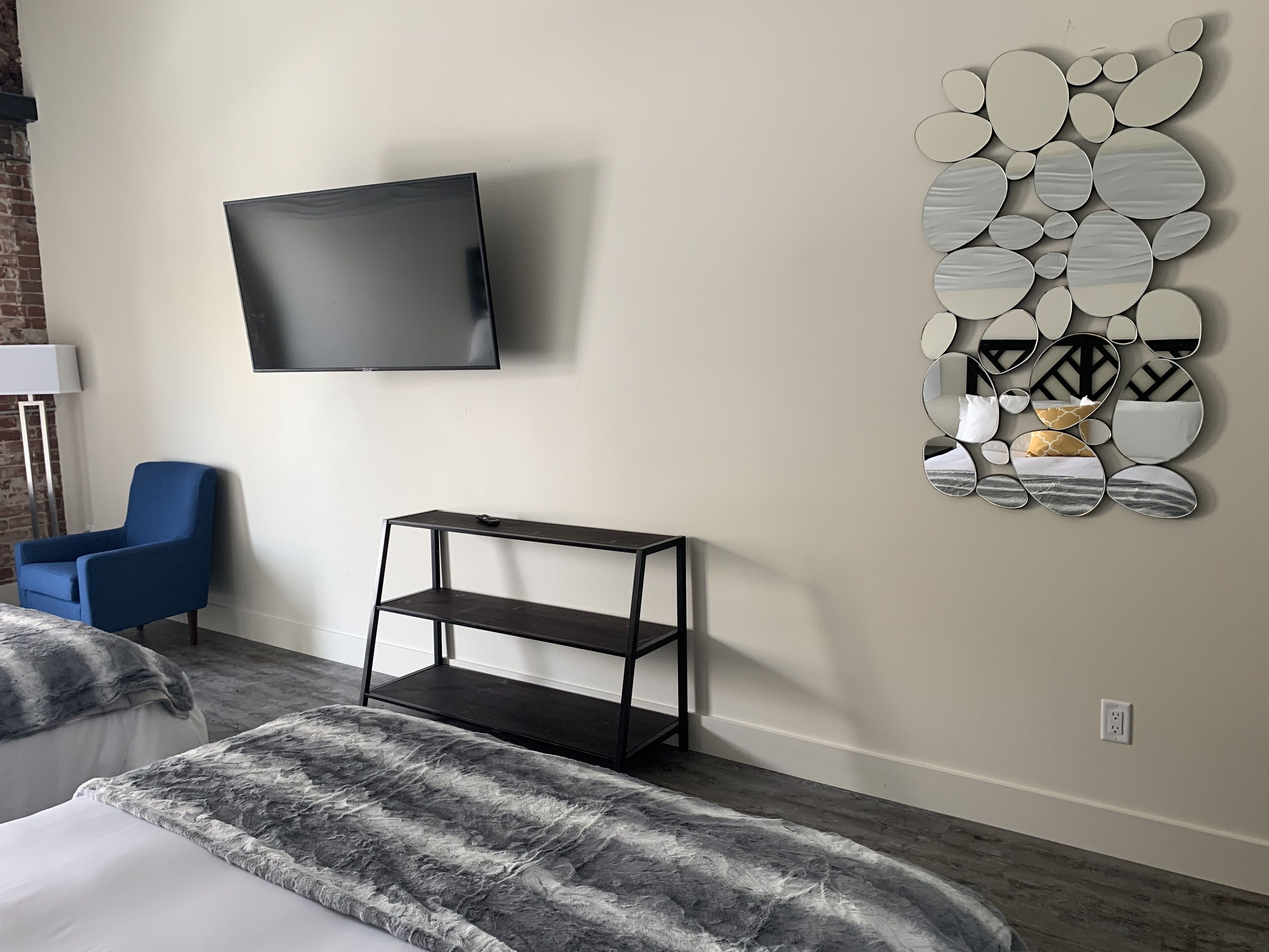 A modern hotel room with a flat-screen TV mounted on the wall, a blue armchair, a black metal shelf, and a decorative mirror wall.