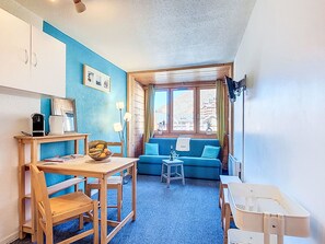 A compact room with a kitchenette, a dining table, a blue sofa, and a window with a view of buildings. - Arcelle 216 by Interhome (Val Thorens)