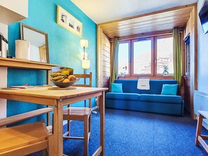 A room with a wooden table, a bowl of fruit, a blue sofa, and a window with curtains. - Arcelle 216 by Interhome (Val Thorens)