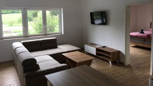Una sala de estar moderna con sofá, mesa de centro, televisor y vistas a un paisaje verde a través de amplios ventanales.