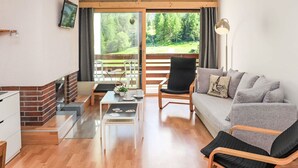 A modern living room with a sofa, a chair, a coffee table, and a TV mounted on the wall. There is a sliding glass door leading to a balcony with a view of trees and a grassy area. - Rosablanche A24 by Interhome (Siviez-Nendaz)