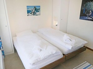 A small, clean room with two single beds, a wooden floor, a small rug, and a painting on the wall.