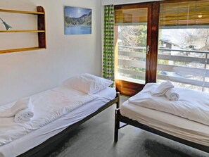 Deux lits simples avec du linge de lit et des serviettes blanches, une étagère en bois, un tableau représentant un paysage, et une fenêtre donnant sur des arbres et un bâtiment.