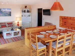 Un salon chaleureux avec une table à manger en bois, dressée pour quatre personnes, un canapé blanc orné de coussins à rayures, un meuble blanc décoratif et un téléviseur fixé au mur.