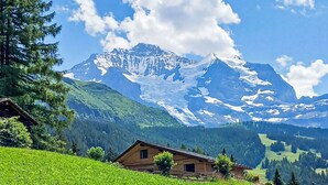 A wooden cabin with a sloped roof, surrounded by lush green fields and a backdrop of snow-capped mountains under a clear blue sky.