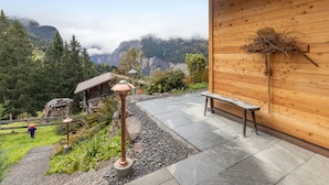 A wooden cabin with a stone patio, a bench, and a view of a mountainous landscape.