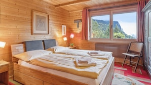 A wooden cabin bedroom with a large bed, a chair, a window with a mountain view, and a radiator.
