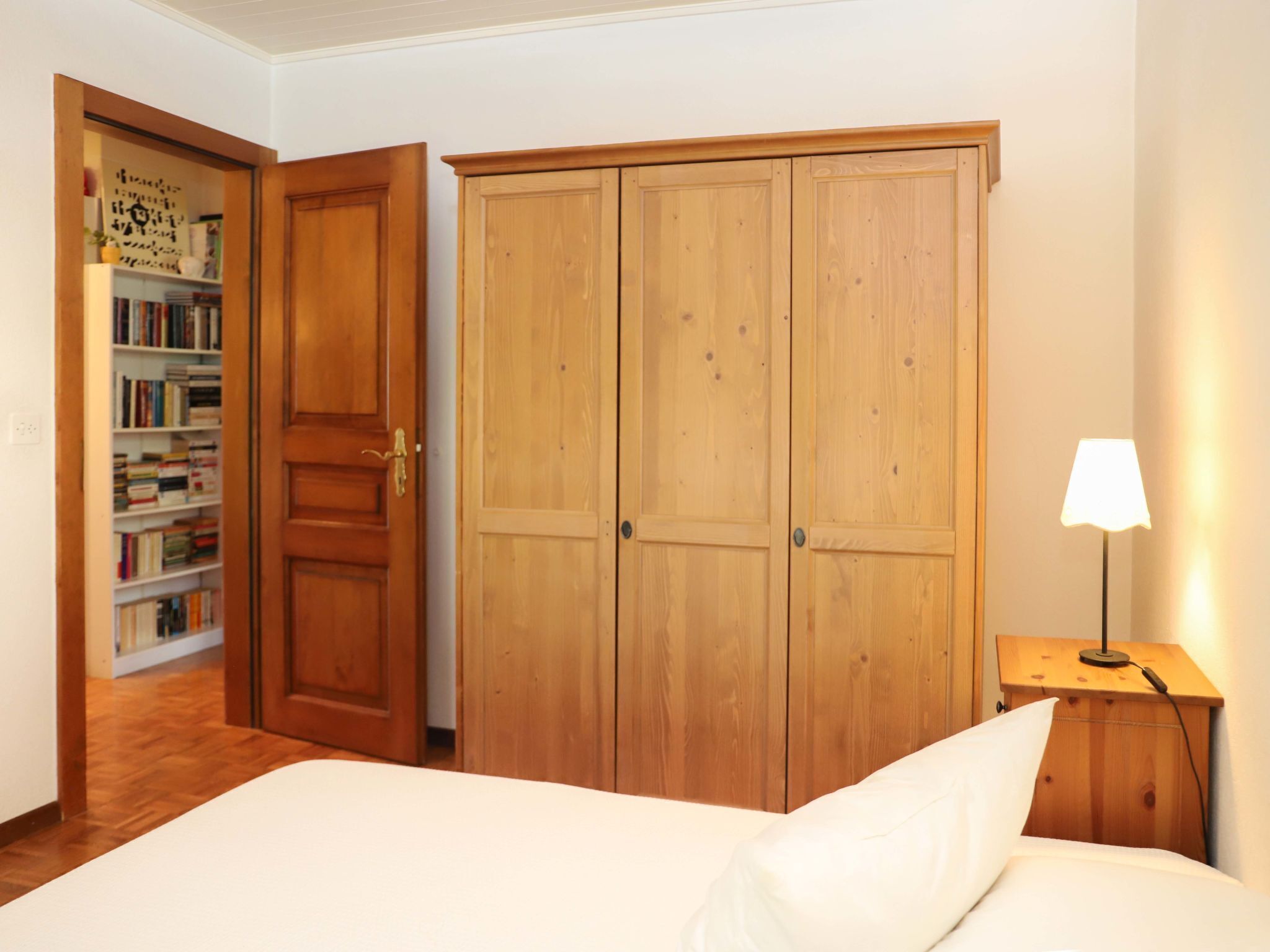 A bedroom with a wooden wardrobe, a bedside table with a lamp, and a bookshelf in the background.