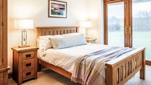 A bedroom with a wooden bed, two bedside tables with lamps, a nightstand with drawers, a framed picture on the wall, and a sliding glass door to the right.