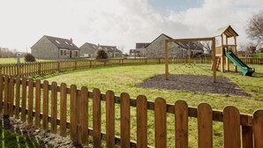 A playground with a slide and climbing net, surrounded by a wooden fence, with houses in the background.