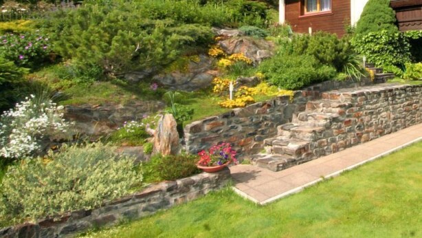 Una casa en una ladera con un muro de contención de piedra, una escalera de madera y diversas plantas y flores.