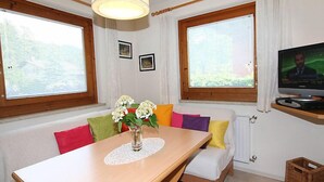 Una habitación con una mesa de comedor de madera, cojines coloridos, un jarrón con flores, un televisor sobre un mueble bajo y ventanas grandes con cortinas.