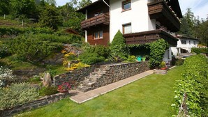 Un chalet de dos pisos con una escalera de piedra que lleva a la entrada, rodeado de exuberante vegetación y un jardín cuidado.