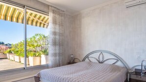 Une chambre avec une grande fenêtre, un lit recouvert d’une couette blanche, une table de chevet et un climatiseur fixé au mur.