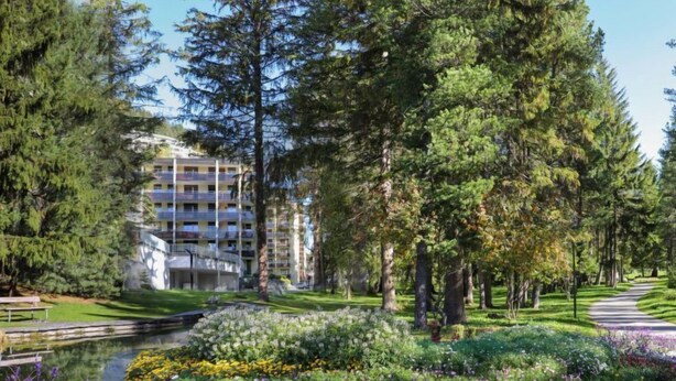 A landscaped garden with a pond, surrounded by dense trees and a multi-story building in the background.