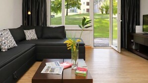 A modern living room with a black sectional sofa, a coffee table with magazines and a vase of flowers, and a television on a wooden stand.