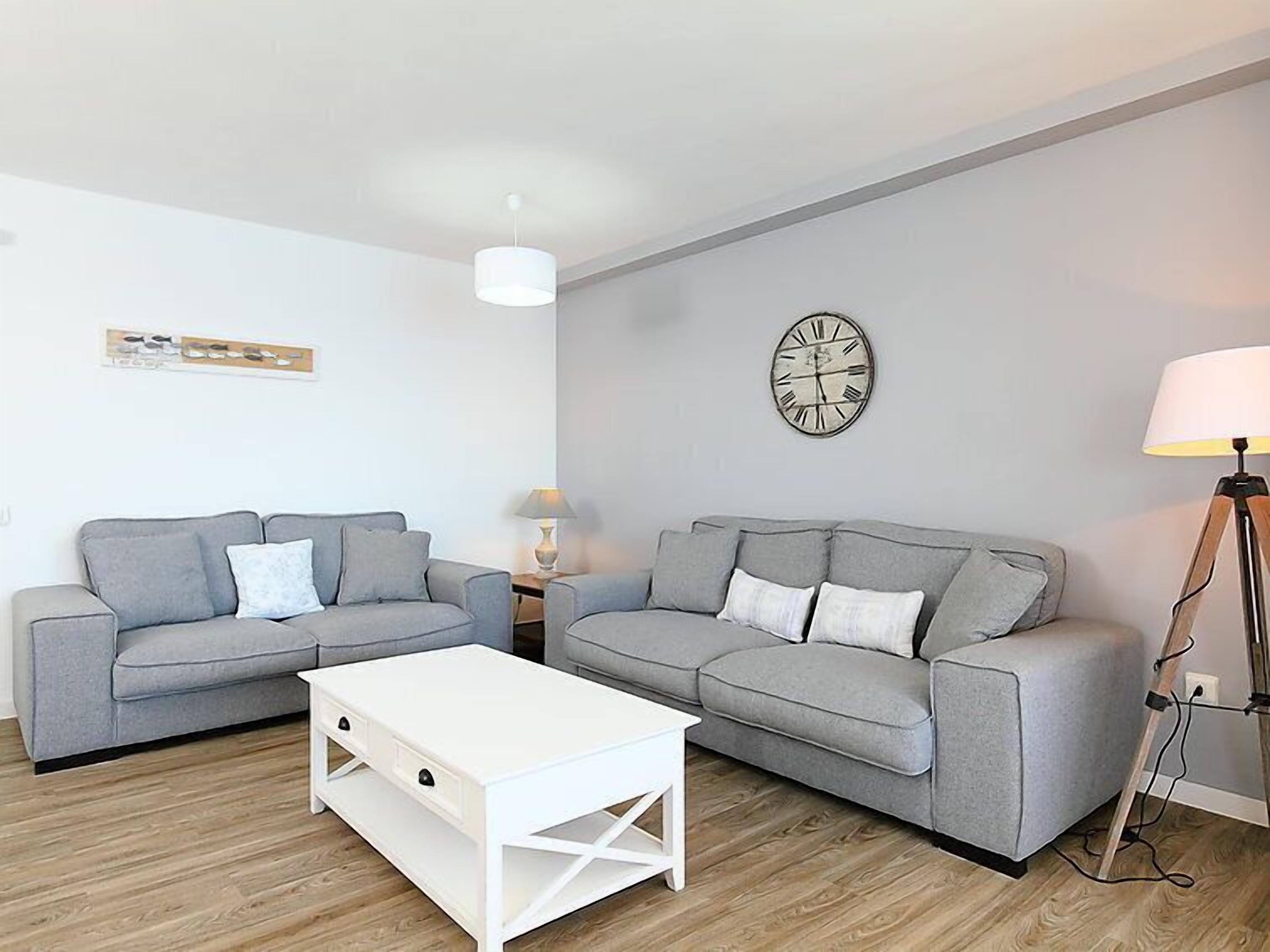 A modern living room with two grey sofas, a white coffee table, a floor lamp, and a wall clock.