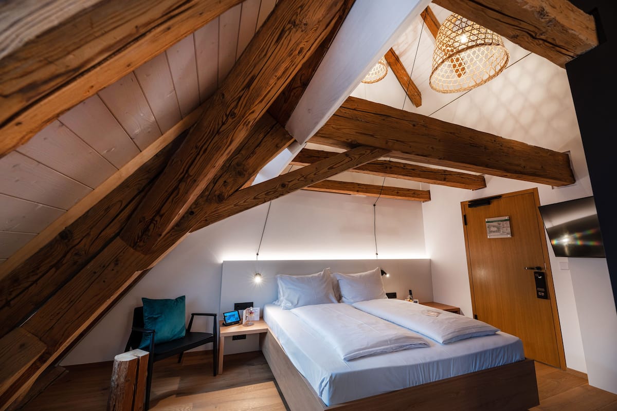 A bedroom with a sloped ceiling, wooden beams, a bed with white bedding, a desk with a laptop, and a hanging light fixture.