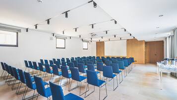 Un salón de conferencias con filas de sillas azules, una mesa blanca con objetos y una puerta de madera al fondo.