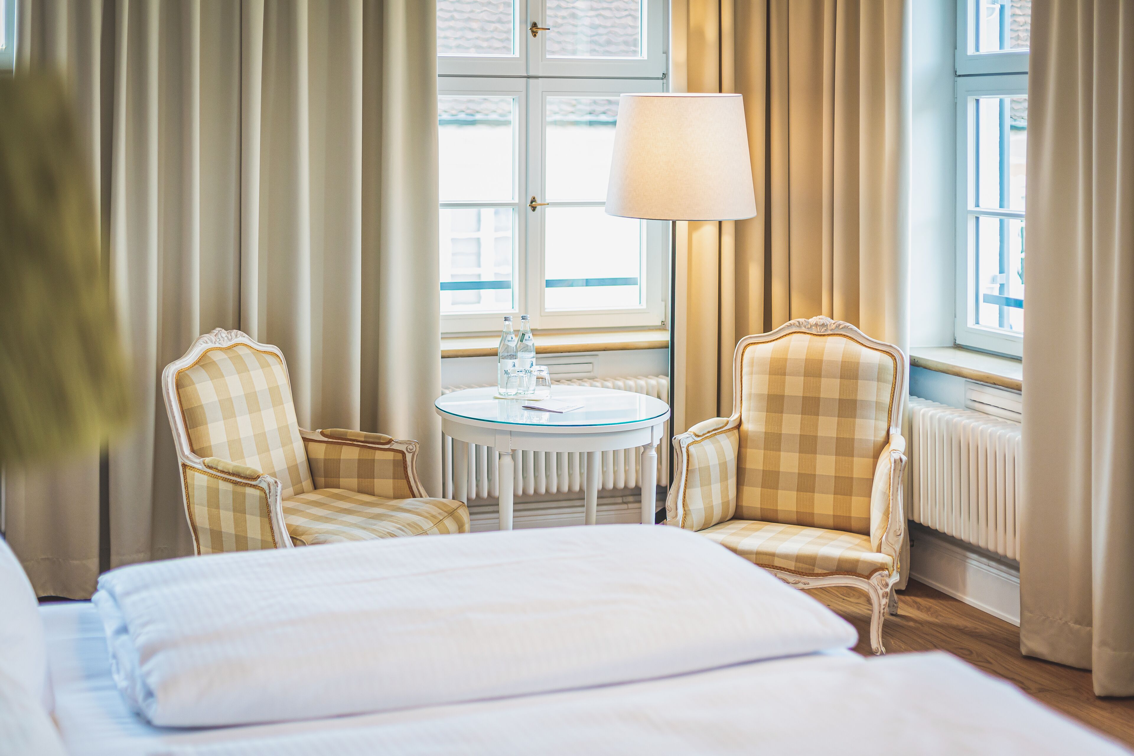 Una habitación de hotel con una cama, dos sillones a cuadros, una mesa redonda con una lámpara y un ventanal con cortinas.