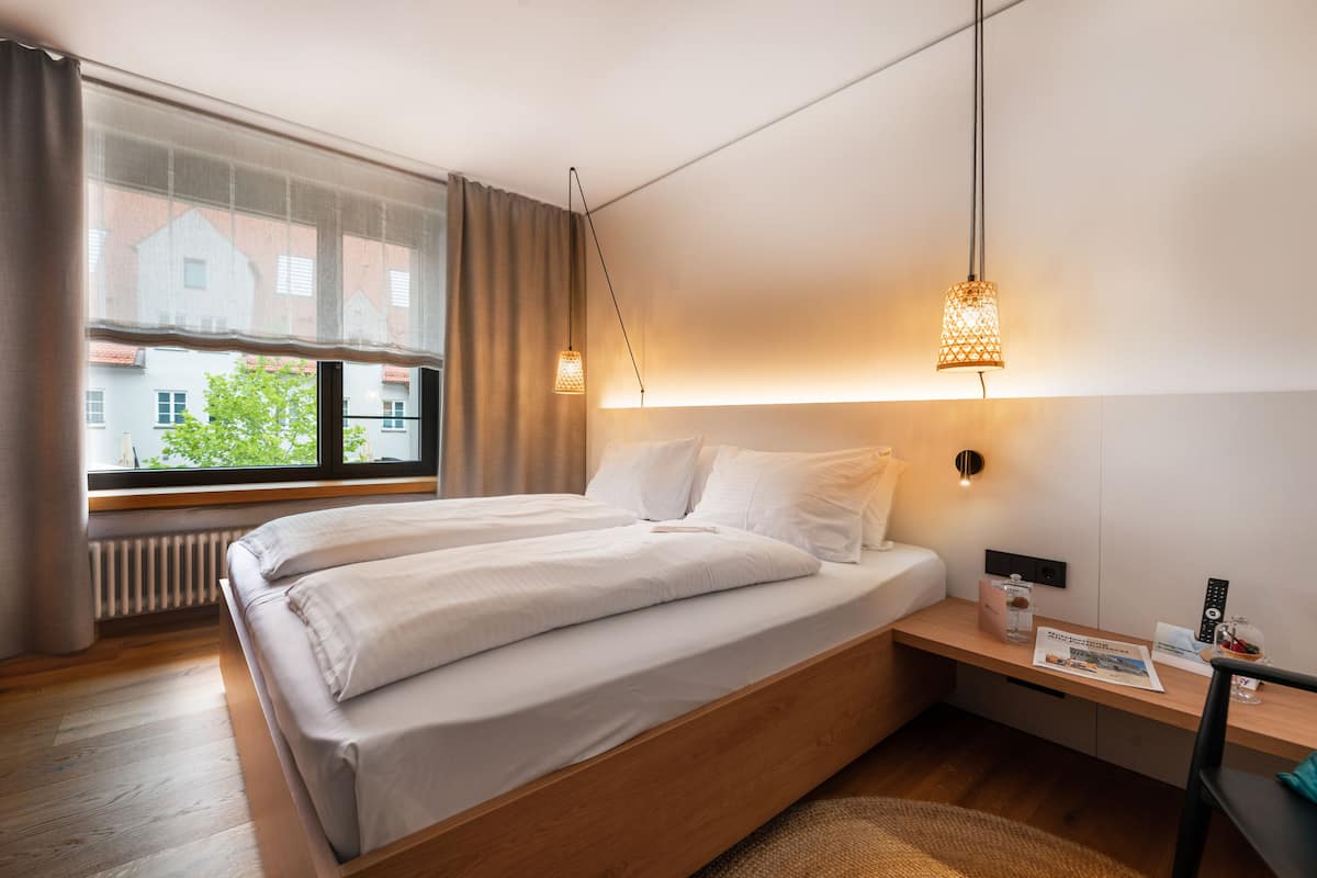 A modern hotel room with a large bed, bedside table, and a view of buildings through the window.