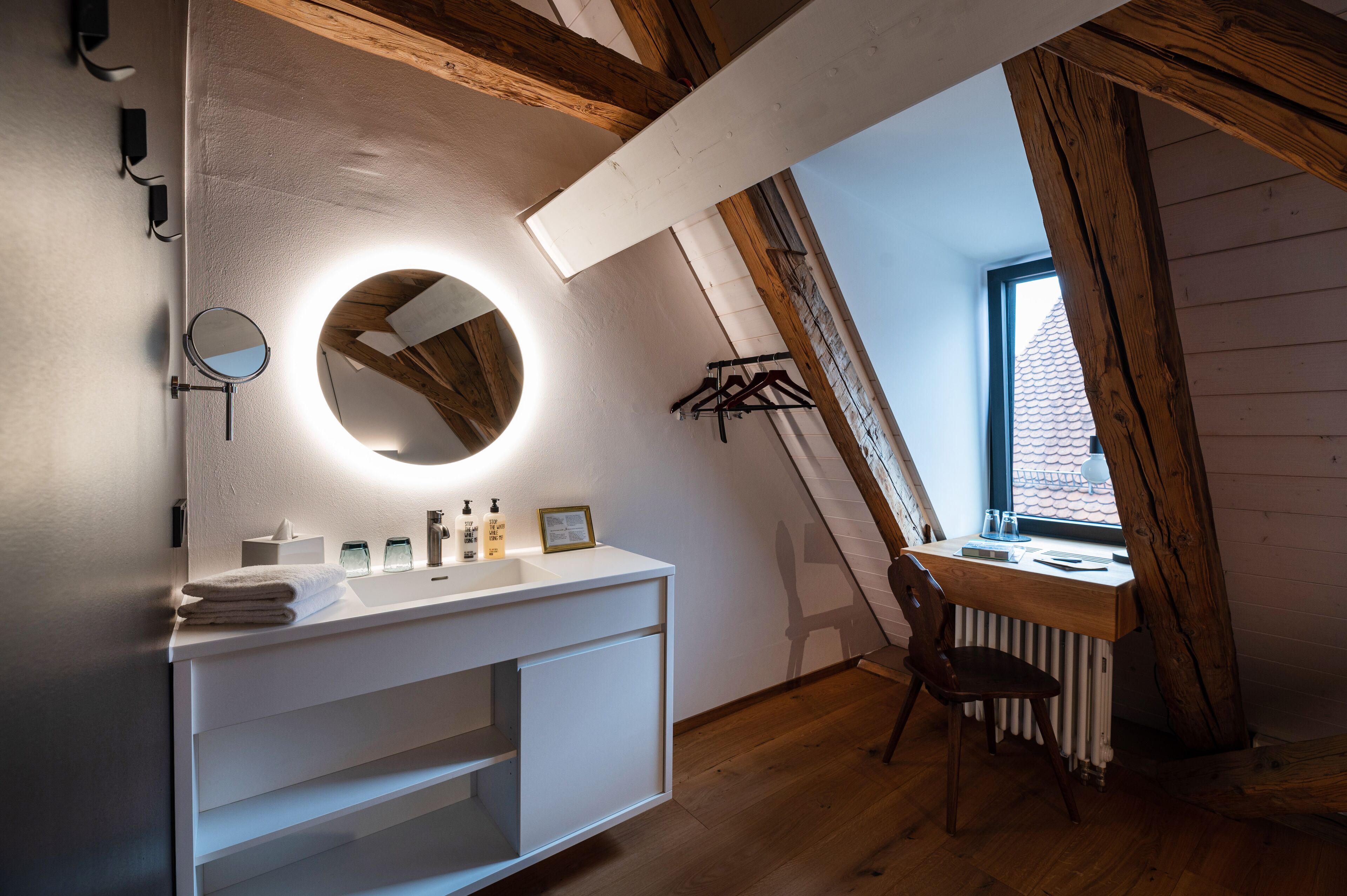 A bathroom with a white vanity, a round mirror, and a window with a view of a tiled roof.