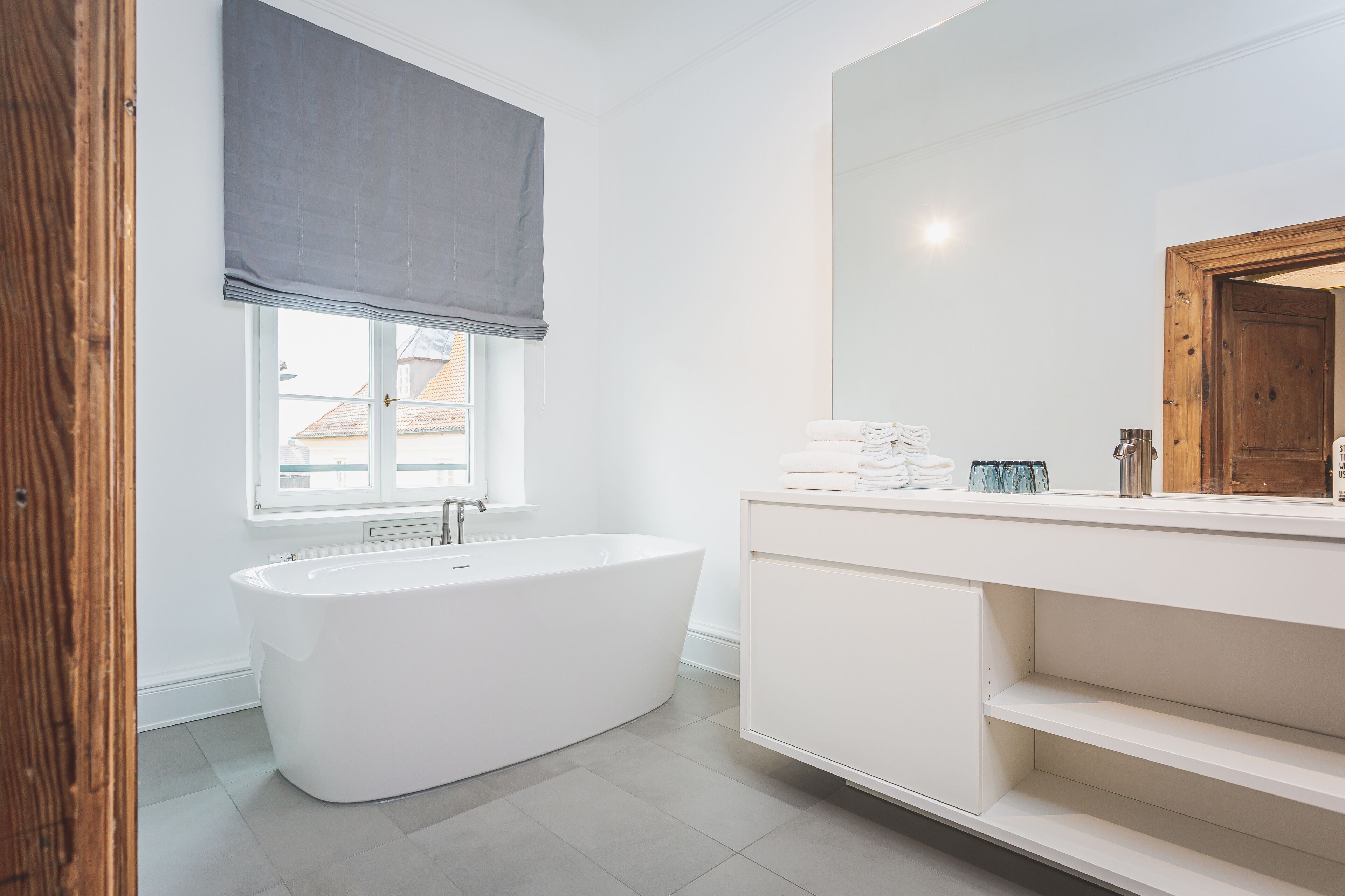 Un baño moderno con una bañera independiente, un espejo grande y una ventana con vistas a edificios.