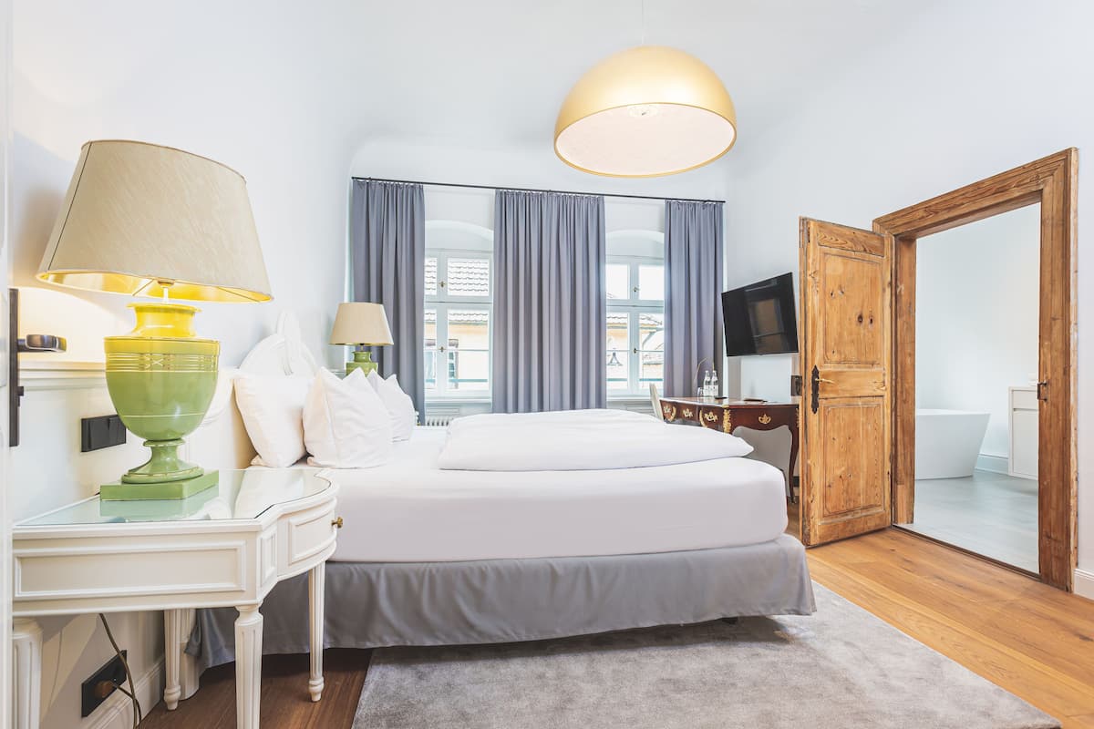 A hotel room with a double bed, two bedside tables with lamps, a TV, a wooden wardrobe, and a view of the outside through large windows.