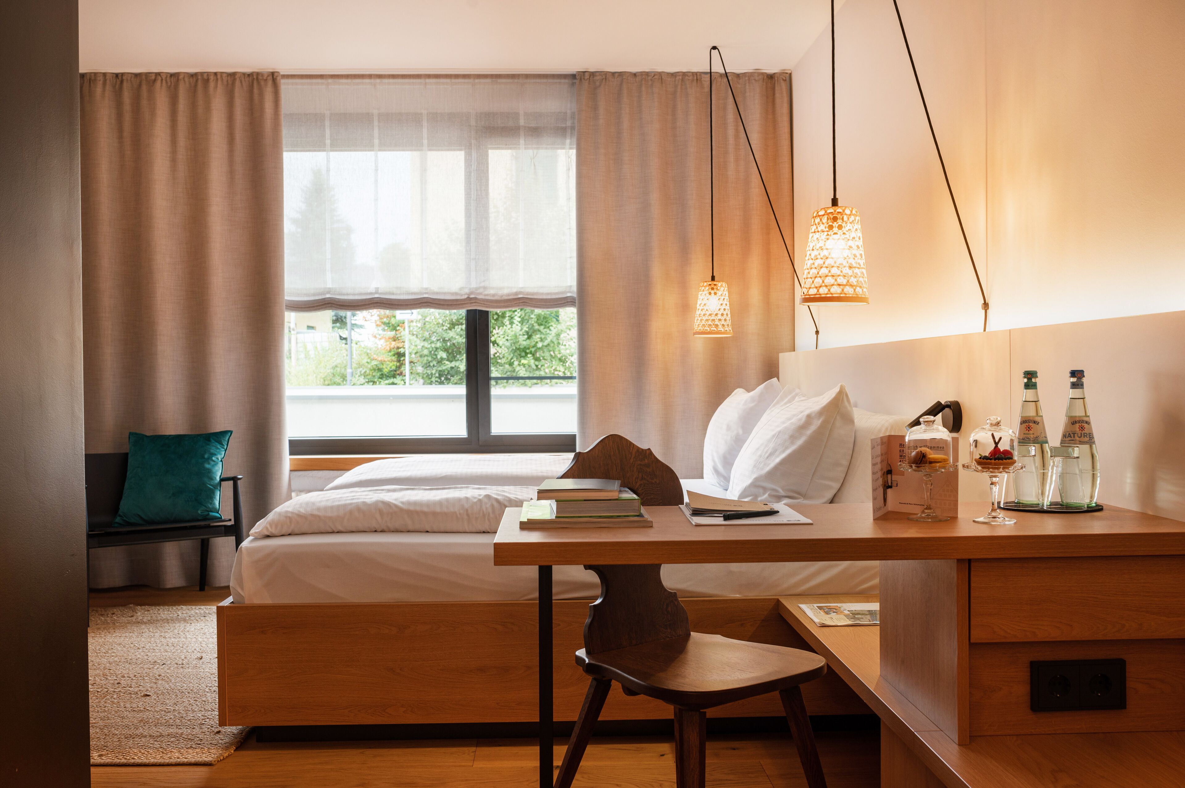 A bedroom with a bed, a desk with books and a lamp, a bench, and a window with curtains.