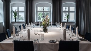 Une salle à manger élégante, avec une longue table dressée pour un repas, de grandes fenêtres ornées de rideaux et des plantes en pot.