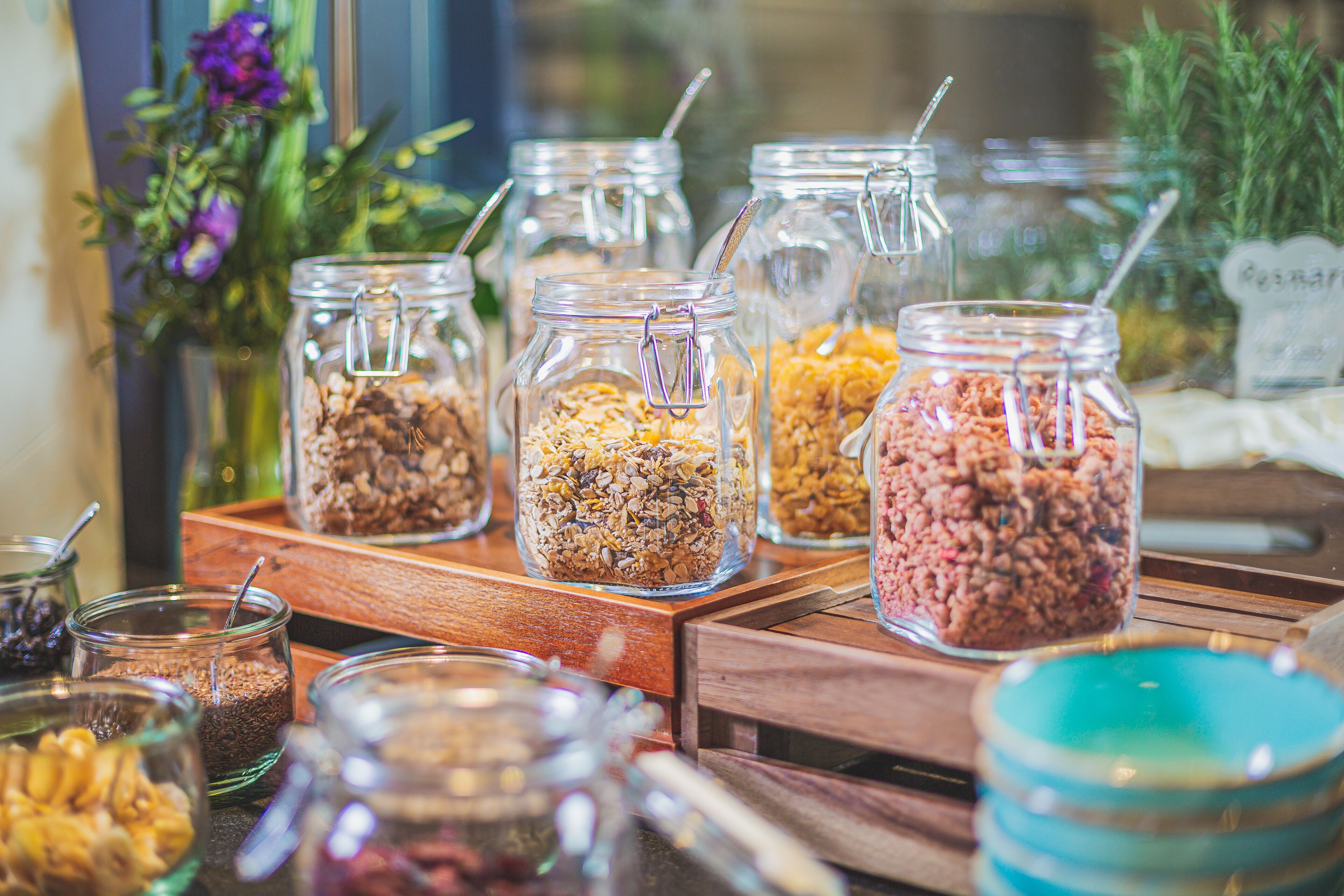 Diversos alimentos en frascos de vidrio sobre una mesa de madera, con un arreglo floral al fondo.
