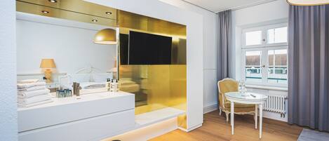 A modern bathroom with a large mirror, a sink, and a toilet. There is a window with curtains and a chair by the window.