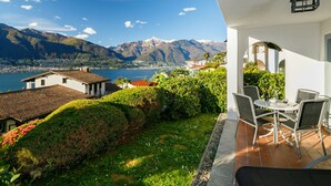Une terrasse offrant une vue sur les montagnes et un lac, entourée de verdure et bordée d’un bâtiment blanc.