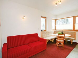 Ein Zimmer mit einer roten Couch, einer Holzbank und einem Esstisch mit Stühlen. Es gibt Fenster mit Holzrahmen und eine Wandleuchte.