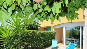 A patio with two blue chairs, a hedge, and a building with a yellow exterior and a glass door.