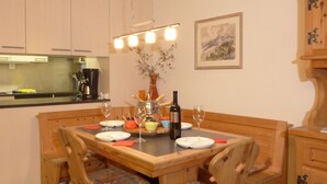 Un comedor con una mesa de madera, sillas y un florero con flores. Se ve una cocina al fondo con armarios y una estufa.