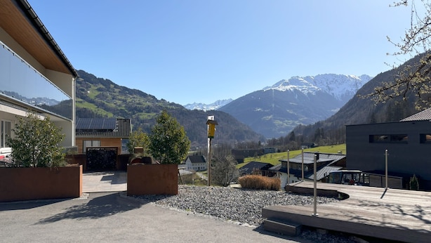 Ein modernes Gebäude mit Solardach, umgeben von einer Schotterfläche und kleinen Bäumen, mit einer Bergkulisse im Hintergrund.