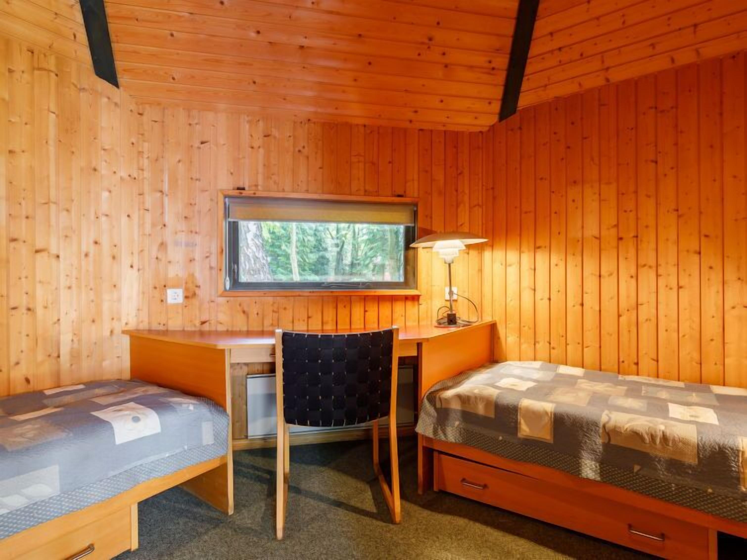 A compact room with wooden walls, a desk, two single beds, a chair, and a window with a view of trees.