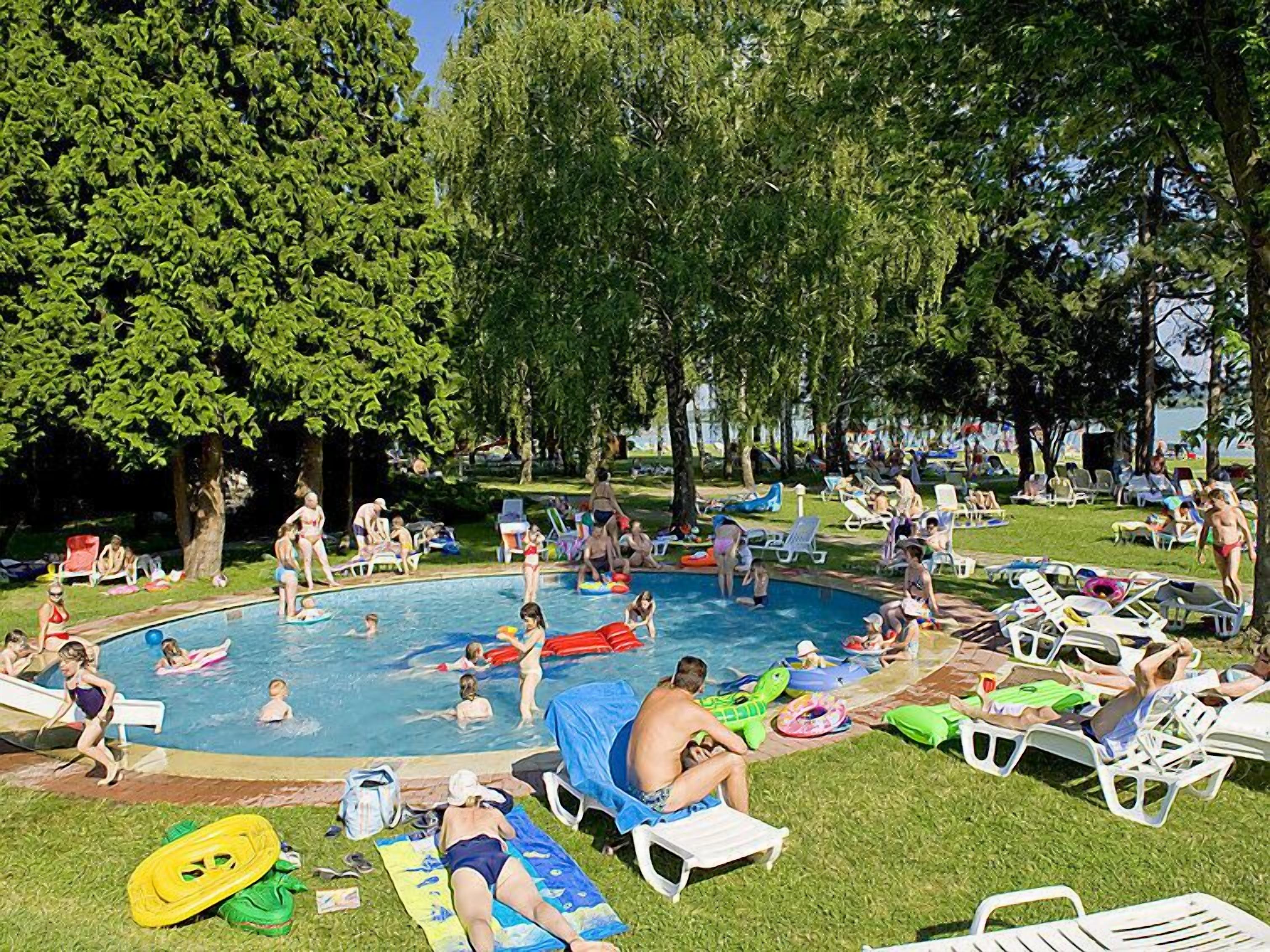 Une piscine entourée de pelouse et d’arbres, où l’on peut voir des gens nager et se détendre au soleil.