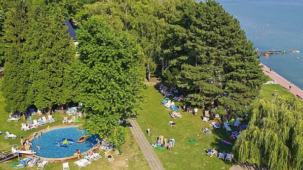 Ein großer Pool im Freien, umgeben von üppigem Grün, mit Blick auf einen See und entfernte Gebäude.