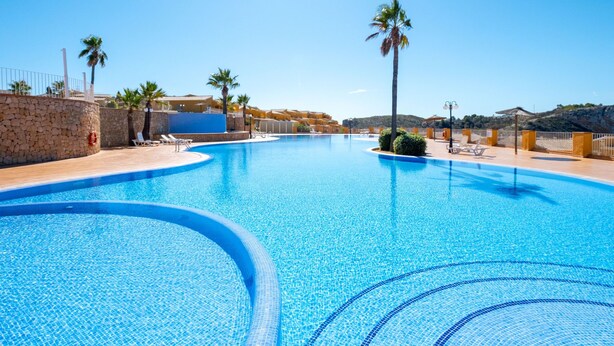 A large, clear blue swimming pool with a stone wall and palm trees, surrounded by a sunny resort setting. - Apartment Pinazo by Interhome (Moraira)