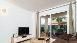 A modern living room with a flat-screen TV on a wooden TV stand, a brown sofa, and a glass door leading to an outdoor patio with a table and chairs. - Apartment Pinazo by Interhome (Moraira)