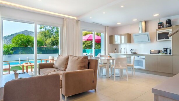 Una sala de estar moderna con sofá, una mesa de comedor y sillas, una cocina con armarios y electrodomésticos blancos, y ventanas amplias con vistas a zonas verdes y una piscina.