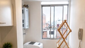 Et lille køkken med vaskemaskine, tørrestativ, vask og et vindue med udsigt over byen.