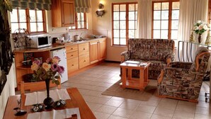 A spacious living area with a wooden dining table, a patterned sofa, and a kitchen with wooden cabinets and a thatched roof. - Otters Den Self Catering Cottages (Fort Nottingham)