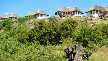 A thatched-roof house with a balcony, surrounded by dense greenery and a large rock formation.
