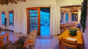 A cozy living room with a sofa, armchair, and coffee table, large windows with curtains, and a view of the outdoors. - Otters Den Self Catering Cottages (Fort Nottingham)