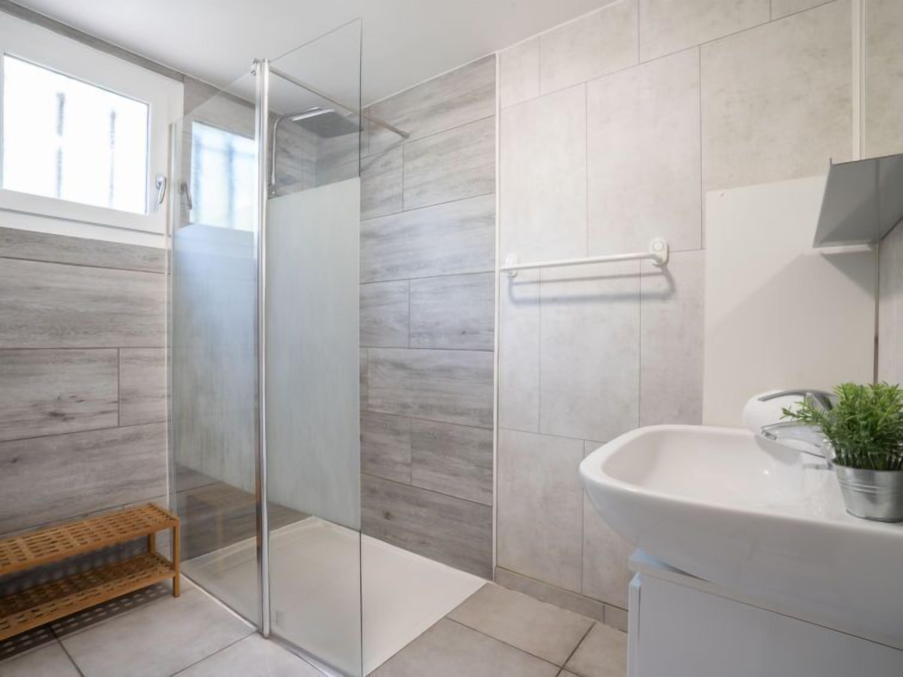 A modern bathroom with a glass shower enclosure, a wooden shelf, and a sink with a potted plant.