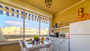 Una cocina compacta con una mesa de comedor, sillas, un refrigerador y un microondas. Una ventana con toldo a rayas ofrece vistas de edificios.