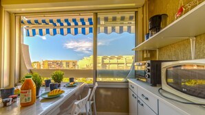 Un pequeño balcón con vistas a edificios, una mesa con comida, un microondas y un estante con utensilios de cocina.